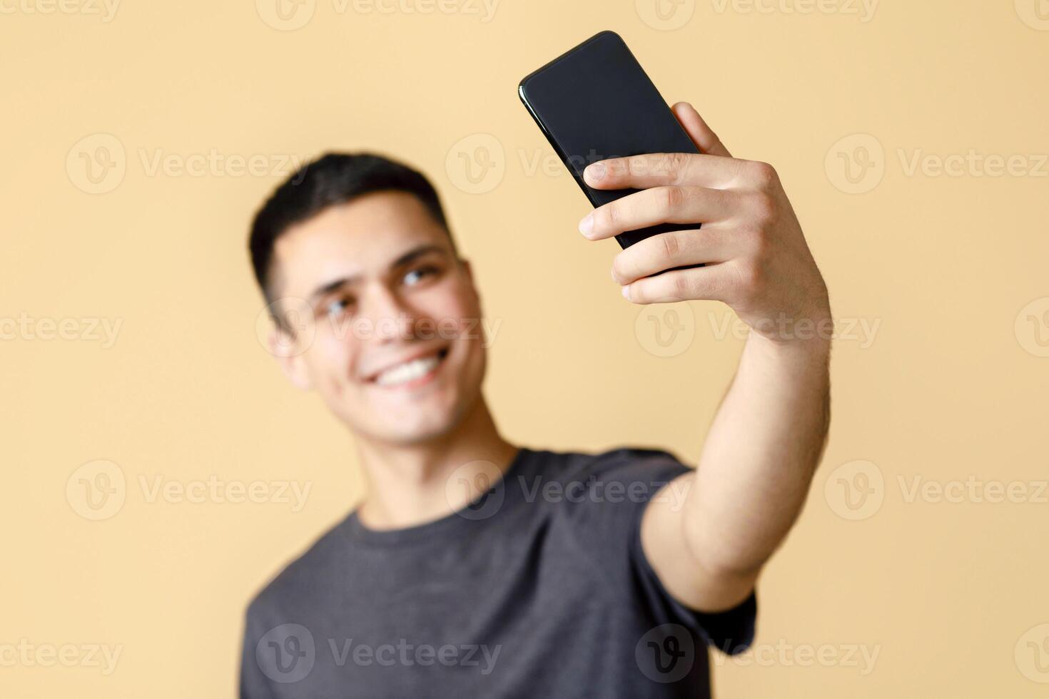 Happy blogger makes streaming or photo for social media. Smiling generation z man in brown t-shirt makes selfie, blurry and focus on smartphone, isolated on beige background, free space