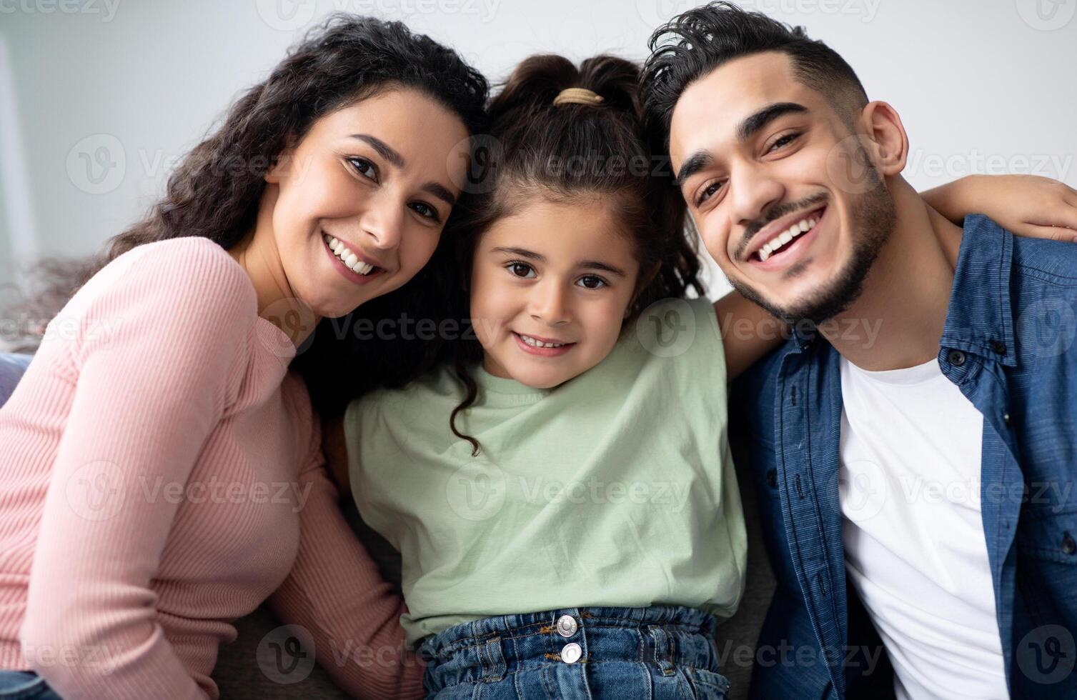 Closeup Portraif Of Happy Arabic Family Of Three With Little Daughter, Cheerful Middle Eastern Parents Posing At Camera While Having Fun With Their Little Child At Home, Relaxing On Couch Together photo