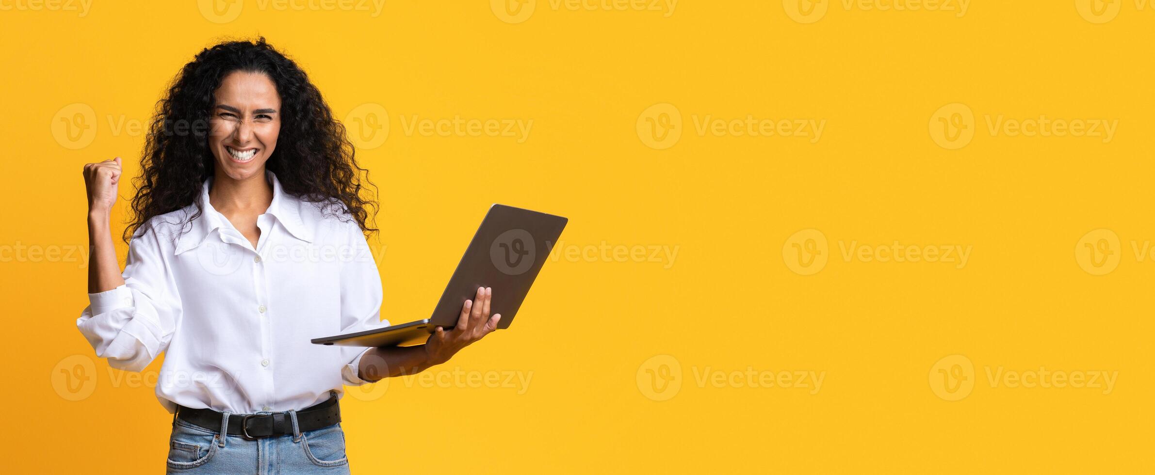 Euphoric Brunette Woman Celebrating Success With Laptop, Raising Clenched Fist, Joyful Millennial Lady Holding Computer, Cheering Victory And Success, Standing On Yellow Studio Background, Copy Space photo
