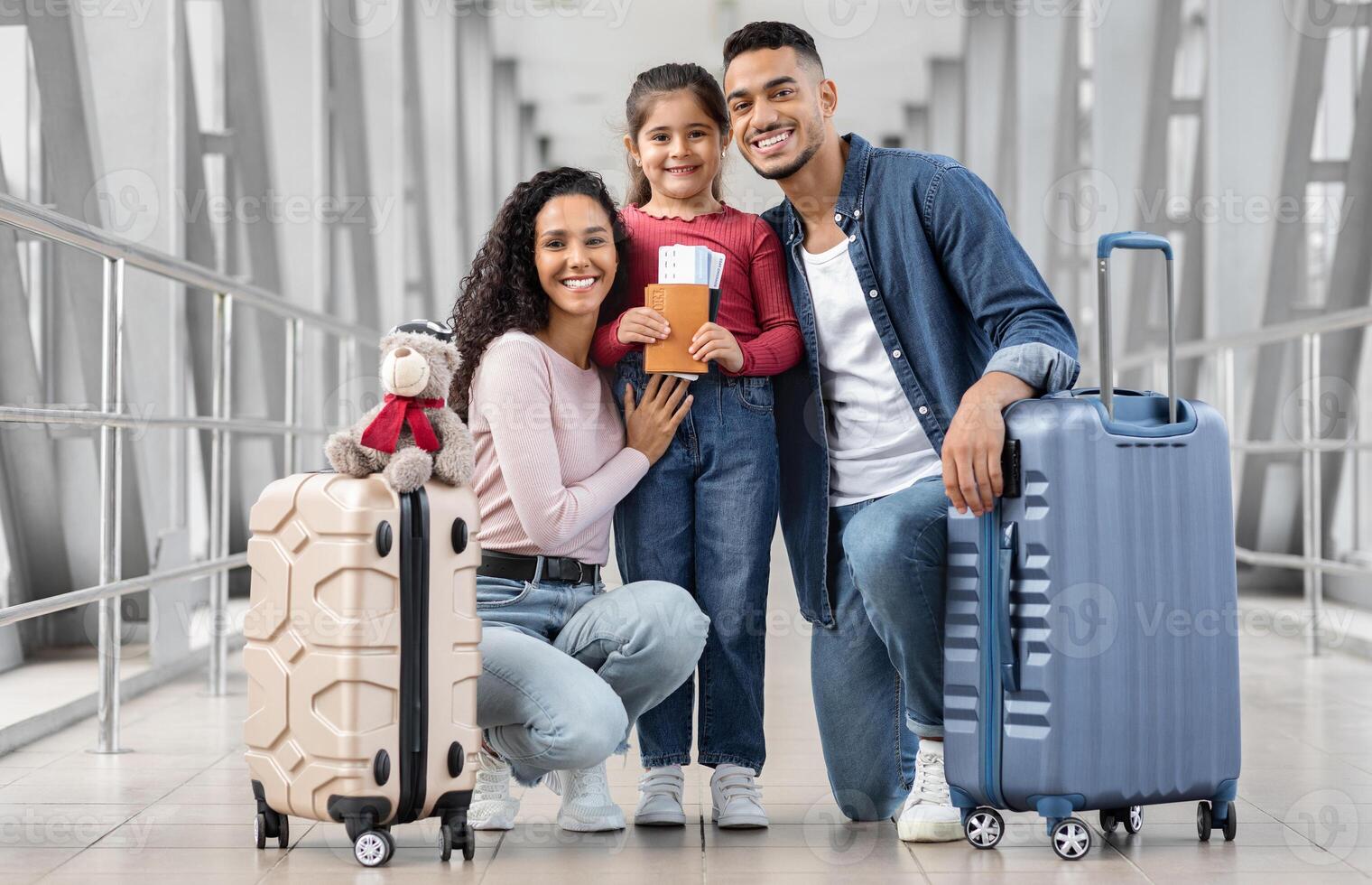 Happy Middle Eastern Family Of Three Posing In Airport Terminal, Cheerful Arab Parents And Their Cute Little Daughter Holding Suitcases, Passports And Tickets And Smiling At Camera, Ready For Travel photo