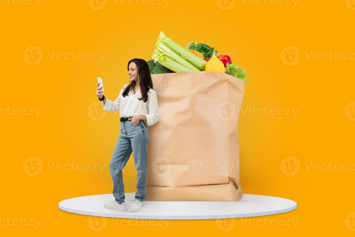 A shopper stands happily beside a large brown paper bag filled with fresh grocery items. They are engaging with their smartphone, possibly ordering groceries for delivery. photo