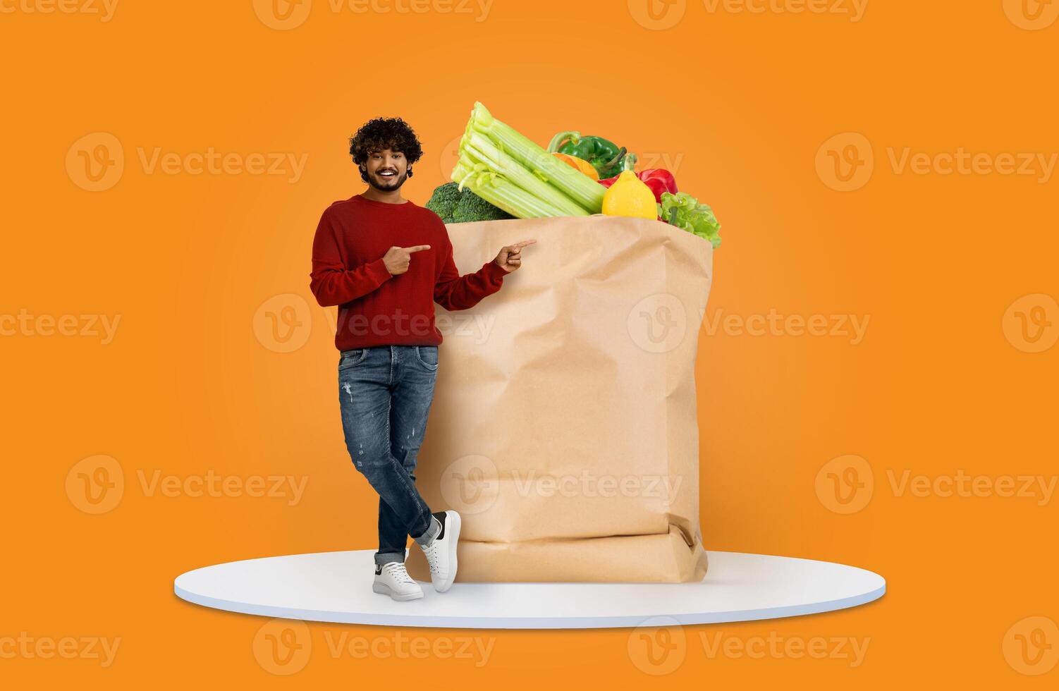 A smiling shopper points at a large paper bag overflowing with fresh vegetables and fruits. The vibrant orange background highlights the new normal of grocery shopping at home. photo