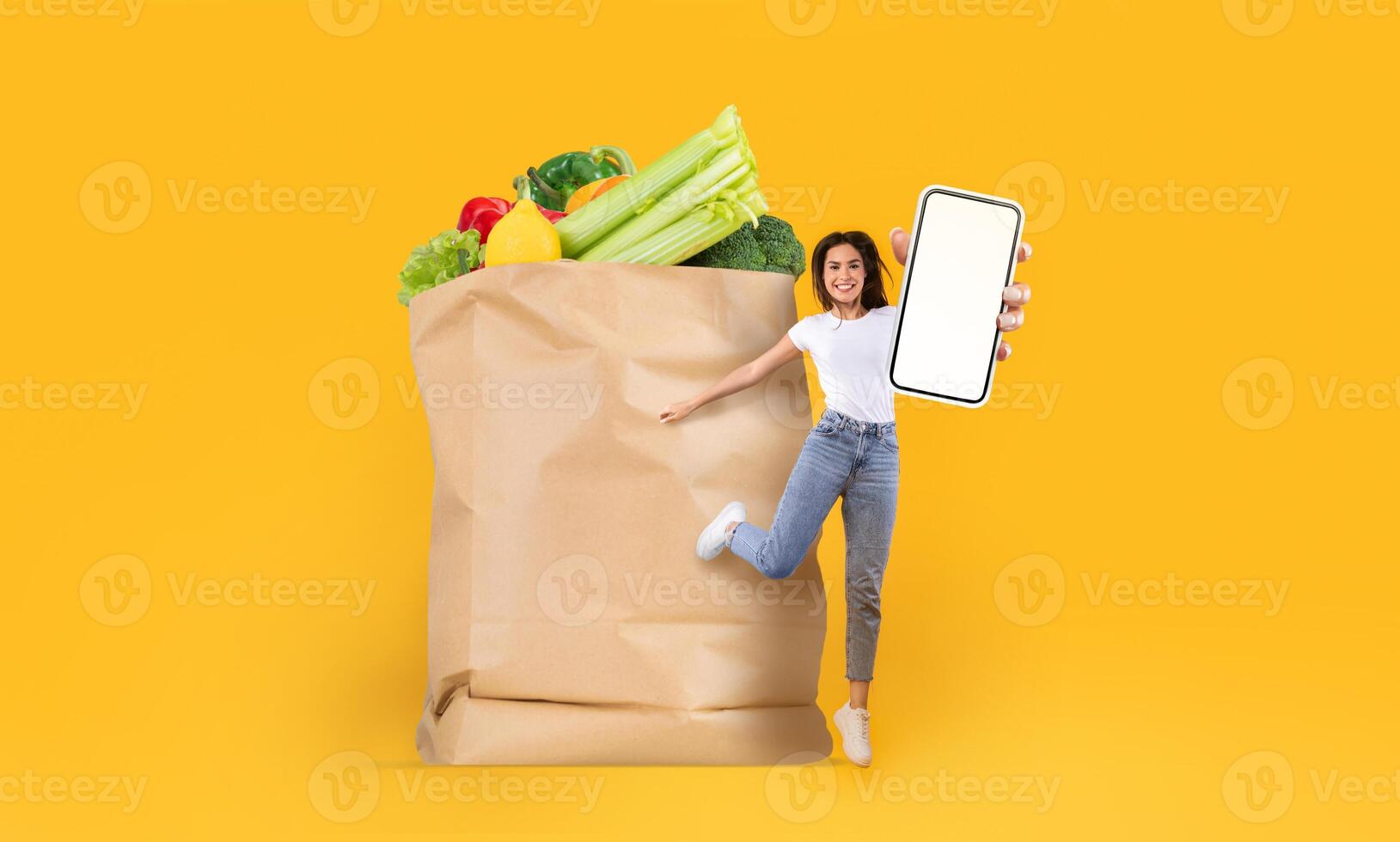 A customer smiles as she stands next to a large paper bag filled with fresh groceries. She holds a smartphone, showcasing the convenience of online shopping. This represents modern grocery purchasing. photo