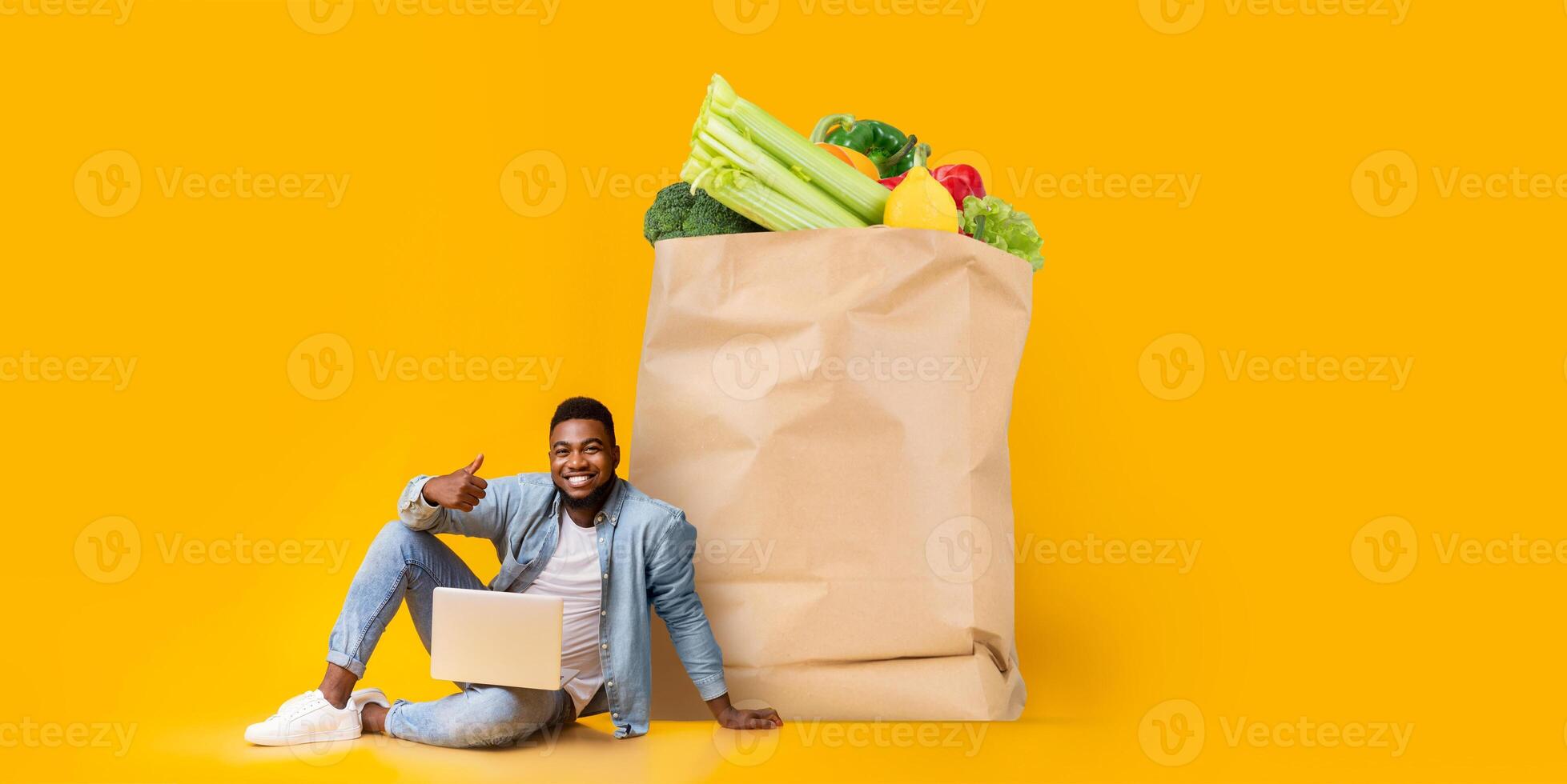 A cheerful customer sits next to a large paper bag filled with fresh groceries while using a laptop to order food. This reflects the new normal of online shopping. photo