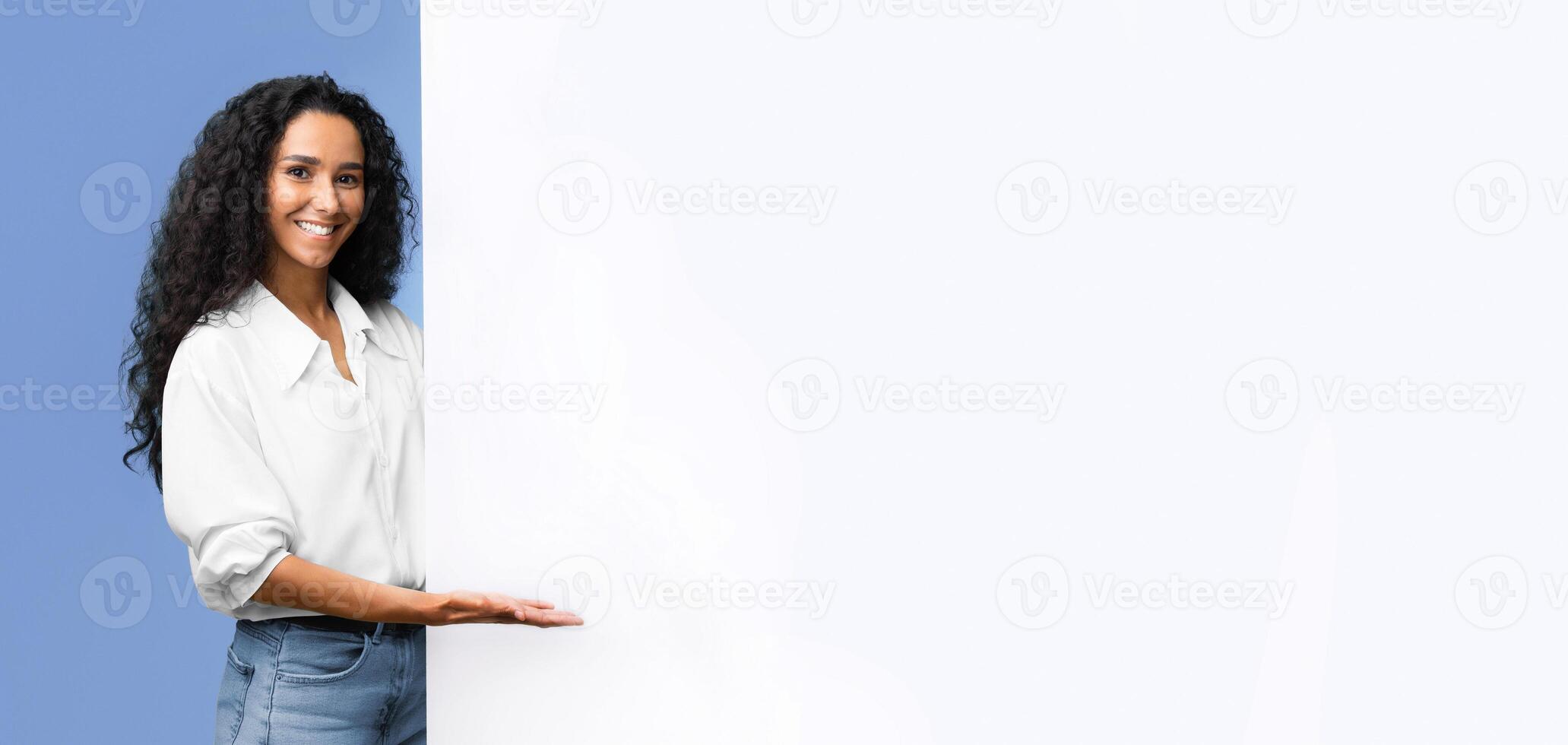 Place For Your Ad. Smiling brunette woman pointing at white advertisement board with open palm, demonstrating copy space for your text or design, standing over yellow studio background, panorama photo