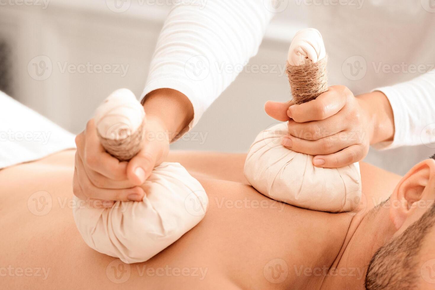 Closeup of compress balls in unrecognizable masseuse hands making herbal massage for handsome bearded man. Middle-aged man spending day at male spa, getting healing herbal massage photo