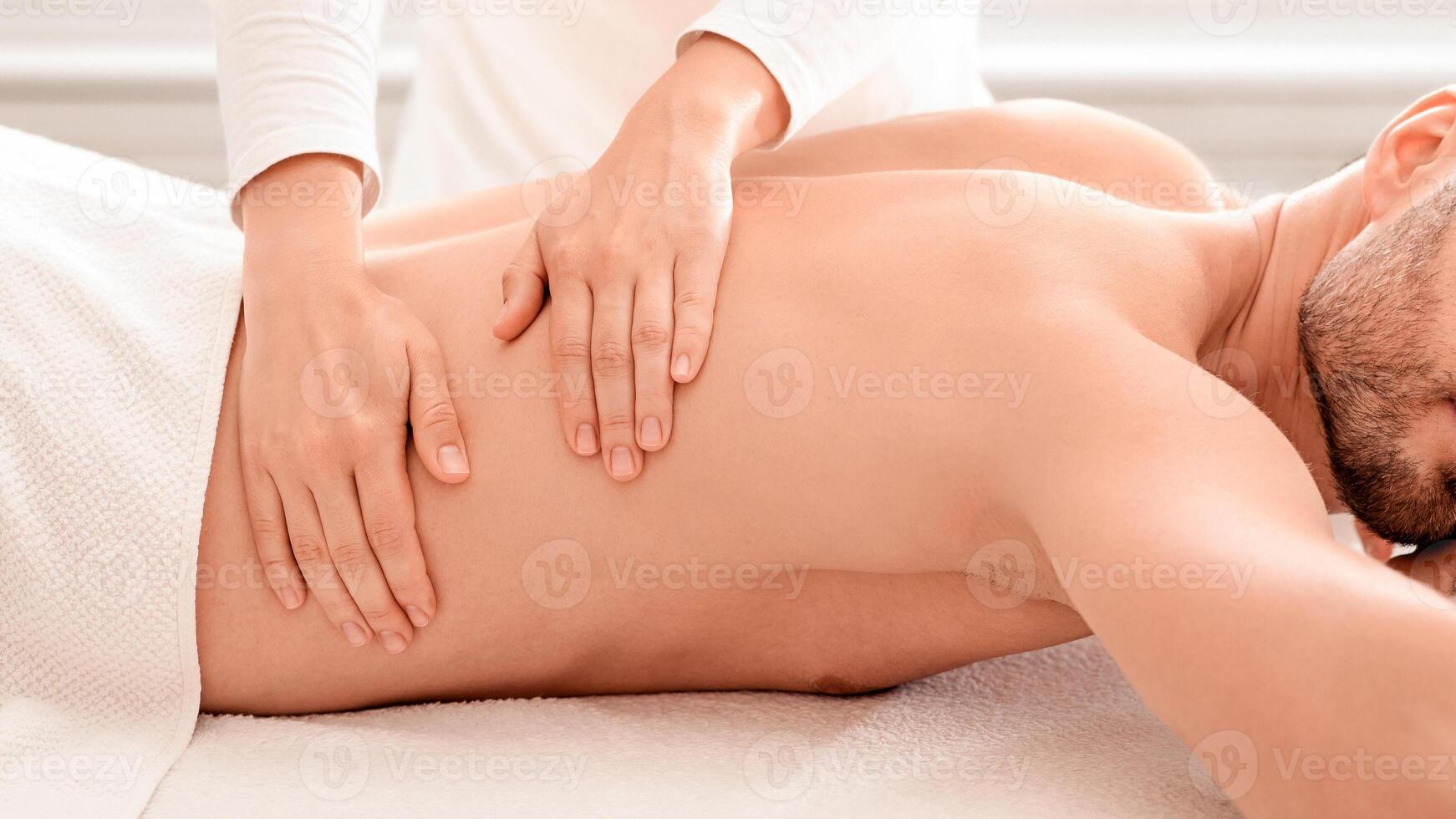 Side view of bearded middle aged man with closed eyes having relaxed body massage at spa salon. Unrecognizable masseuse rubbing sleeping handsome man back. Stress relief concept photo