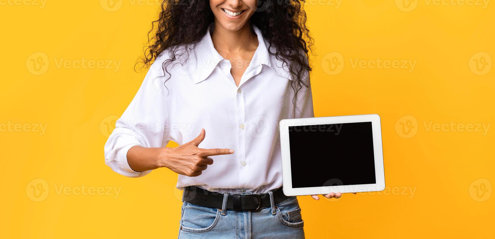 Check This. Positive Young Woman Holding And Pointing At Digital Tablet With Black Blank Screen, Posing Over Yellow Studio Background, Demonstrating Free Space For Your Design, Mockup Image photo