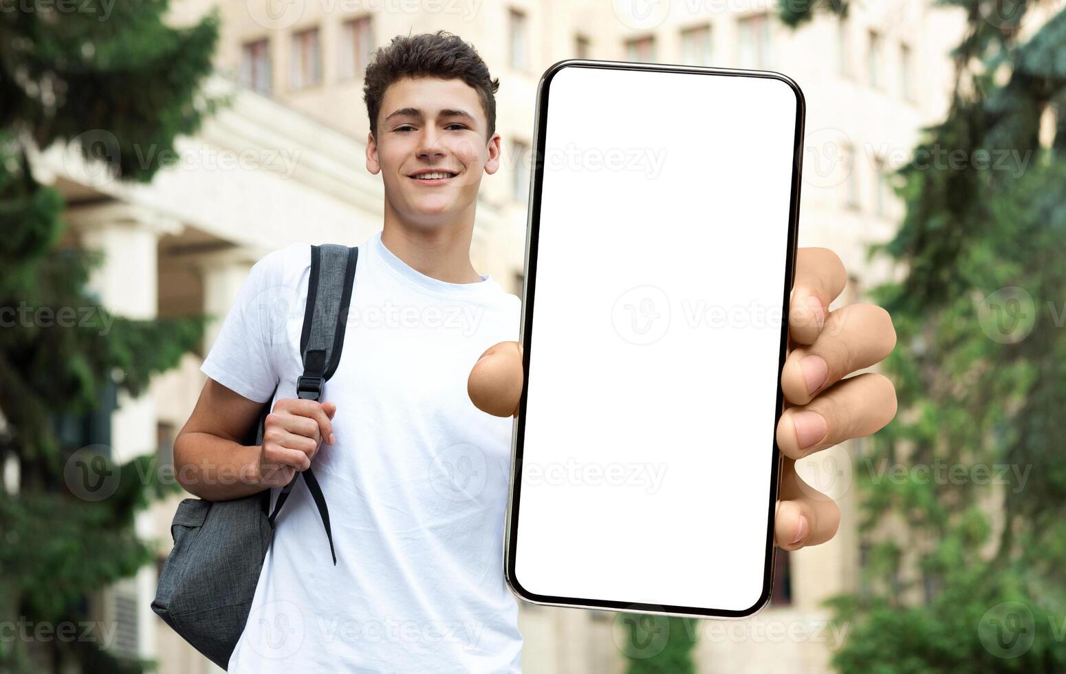 Young Male Student Showing Blank Mockup Screen Holding Big Phone In Hand Advertising Educational Application For Smartphone Standing Outdoors Near College Building, Showing Gadget Close Up To Camera photo
