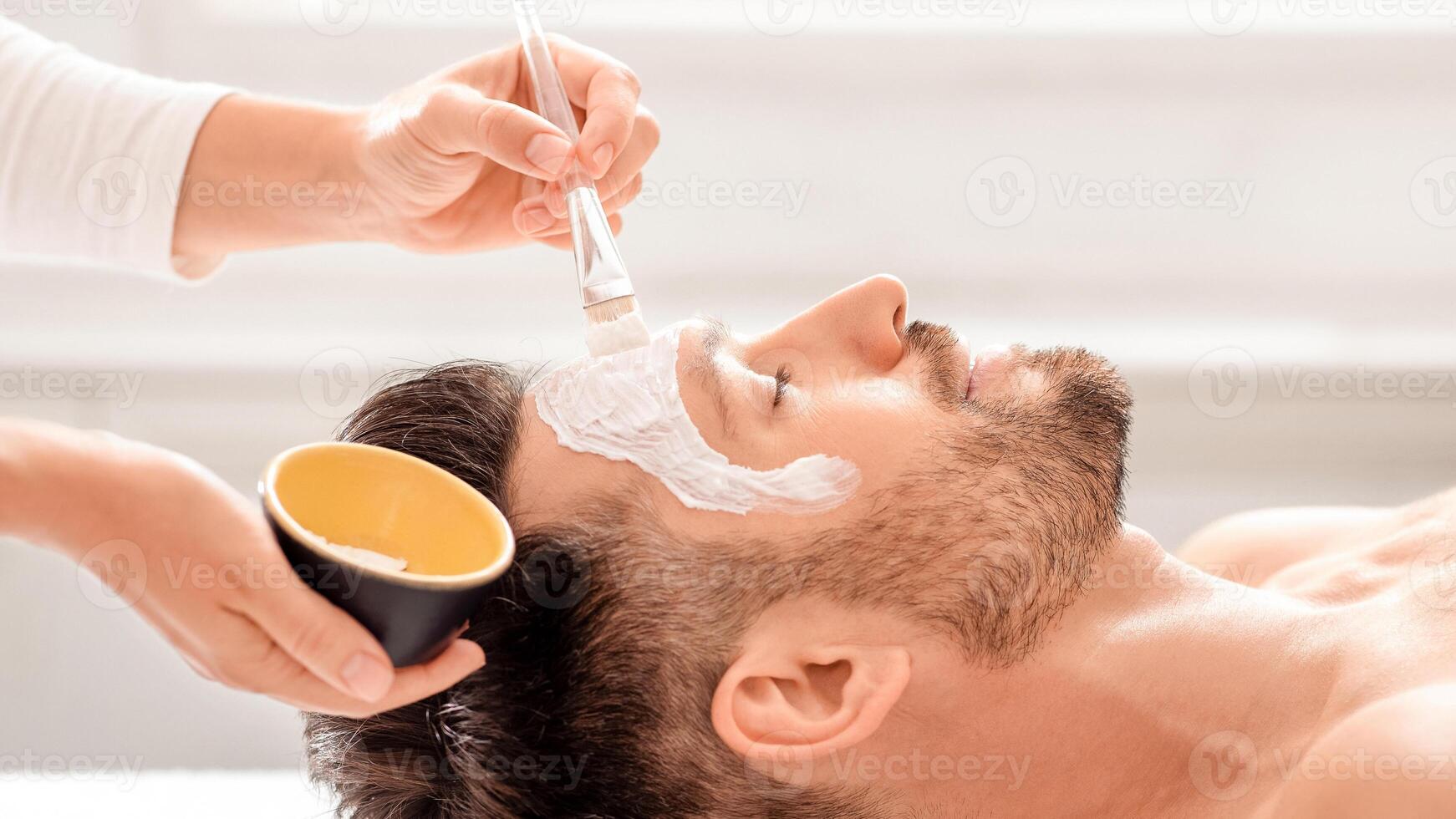 Side view of bearded man getting face treatment at modern spa salon. Female attendant applying nourishing face mask on sleeping middle aged man face. Male cosmetology and face care concept photo