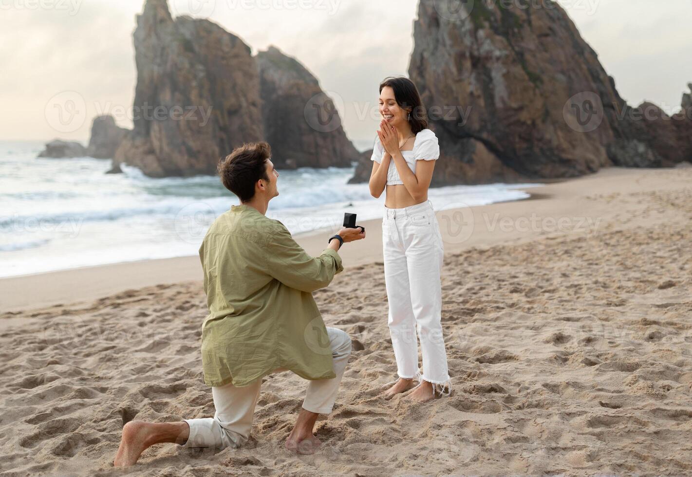 Full length shot of loving man with engagement ring proposing to surprised lady, guy standing on knee on ocean beach, free space. Love is in the air photo