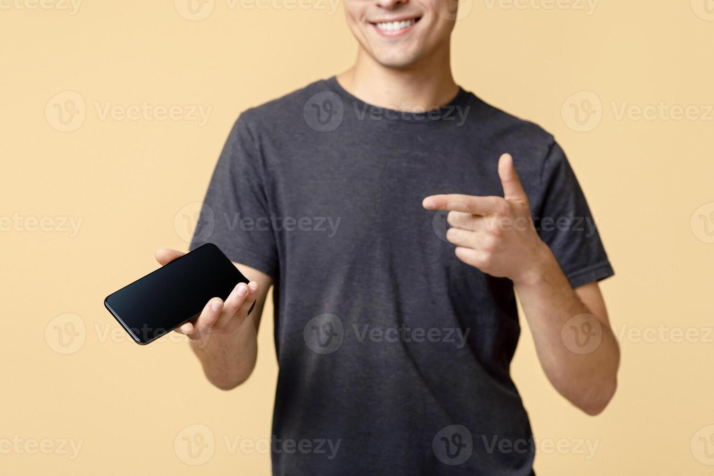 Good offer received on mobile phone. Friendly cute young man in brown t-shirt and glasses, points finger at smartphone with blank screen, isolated on beige background, studio shot, free space photo