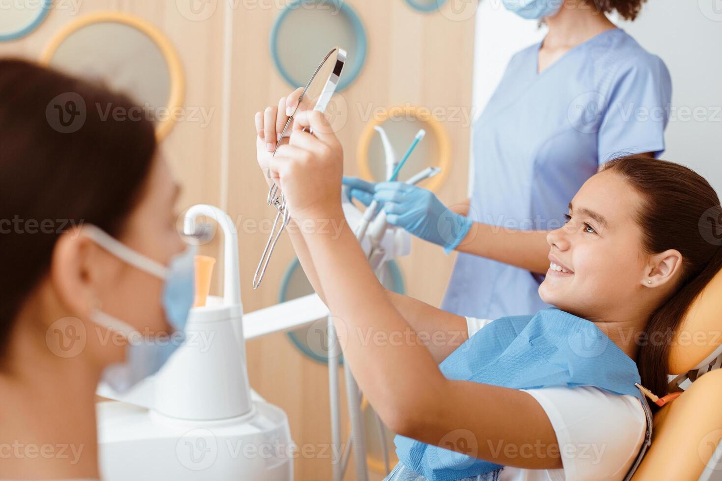 Satisfied dentist clinic patient look at her perfect smile in mirror after treatment in office with medical equipment in background and nurse in white coat, protective mask and rubber gloves, cropped photo