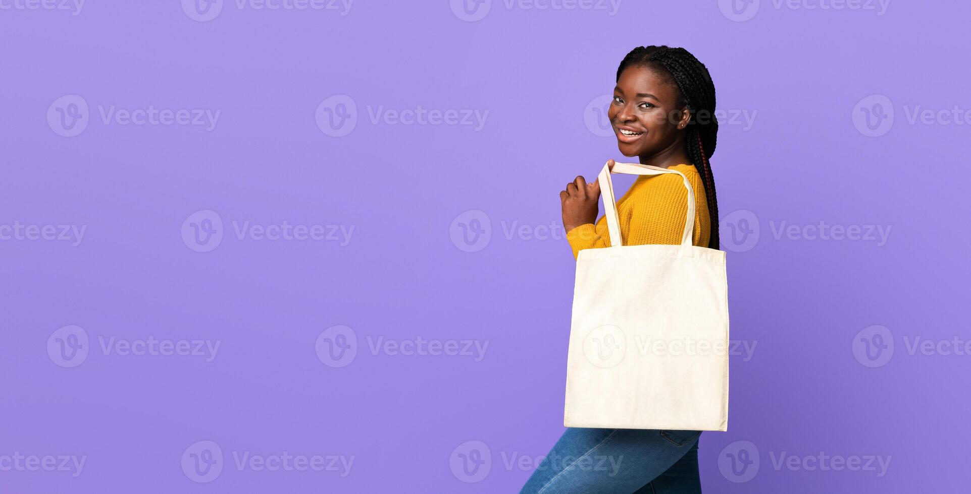Joyful African American Female Posing With Blank Eco Tote Canvas Bag With Mockup For Your Design Or Logo, Positive Black Woman With Braids Standing Over Purple Studio Background, Smiling At Camera photo