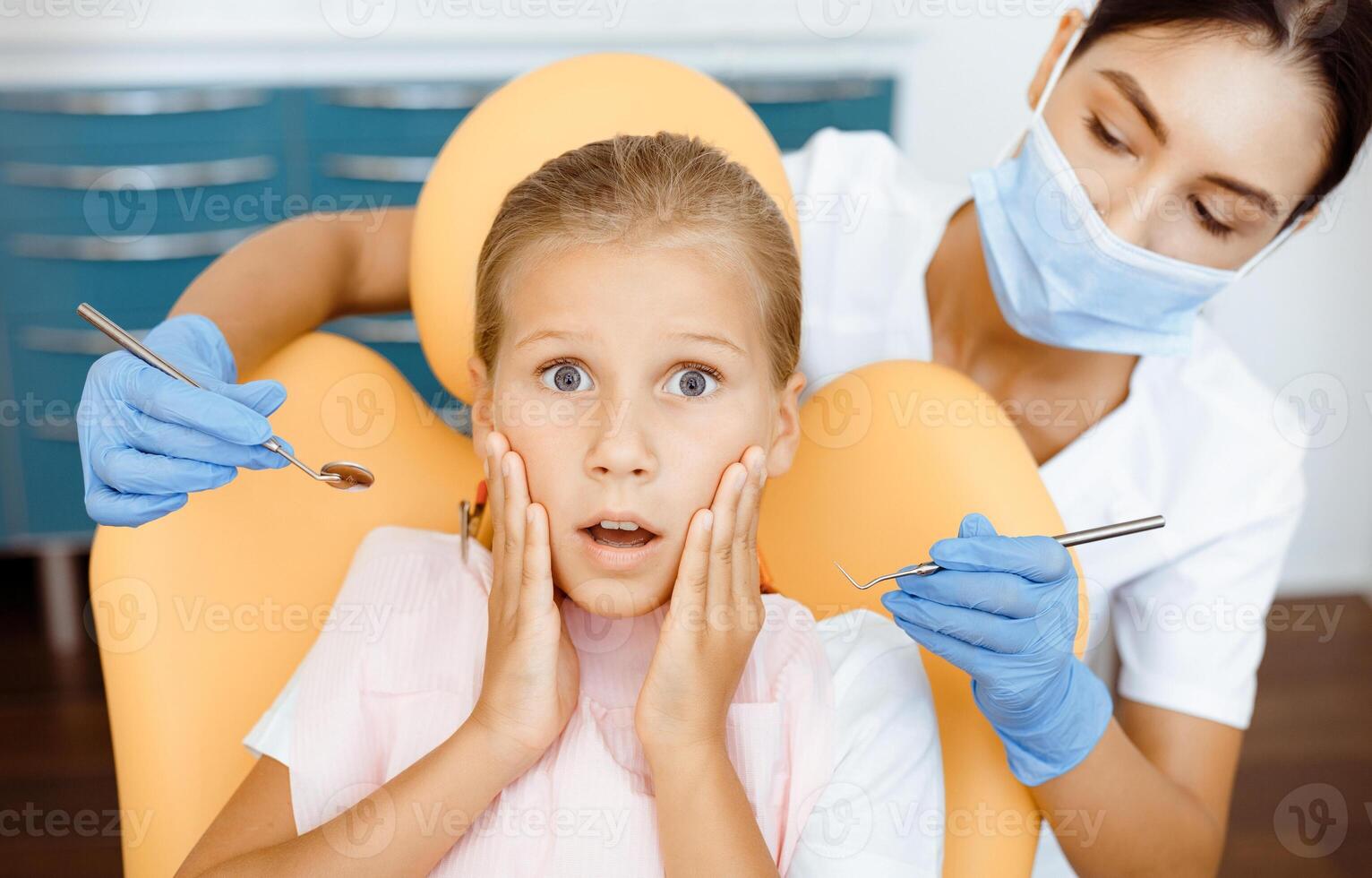 Doctor work with frightened little child in modern office. Frightened girl with open eyes and mouth sits in chair, woman dentist in protective mask and gloves with tools in hands going to treat baby photo