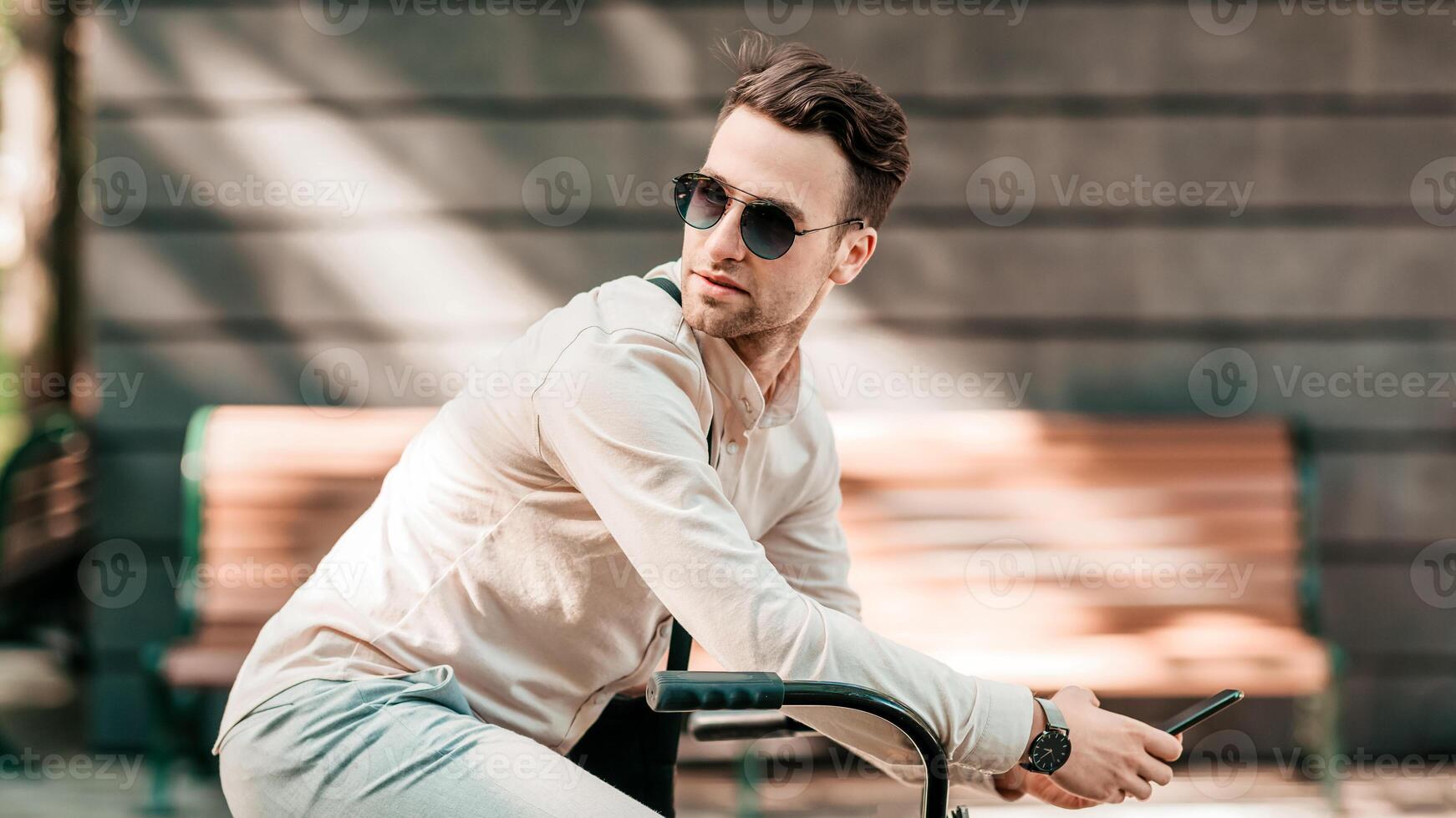 A young man casually rides his bike along a city street. He wears sunglasses and looks engaged while using his smartphone. The sun casts playful shadows around him, enhancing the lively atmosphere. photo