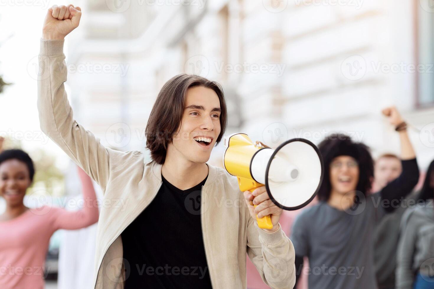 Charismatic guy activist with megaphone on the street, chanting slogans and raising fist up, young caucasian man leading international group of demonstrators, striking against lockdown rules photo