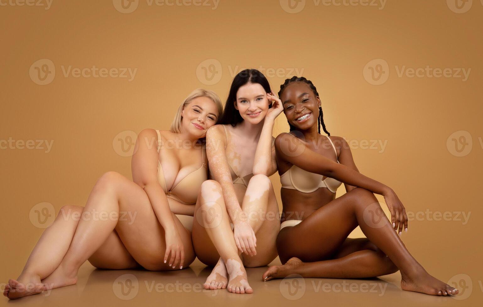 Body Positive. Three attractive multicultural women in lingerie with different skin and body type sitting on floor, posing together in studio photo