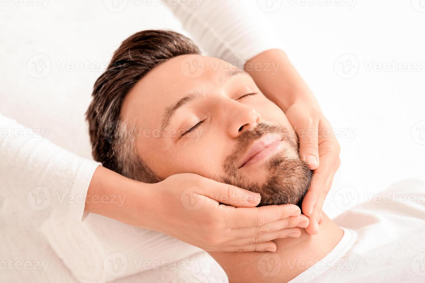 Spa therapist making relaxing massage for handsome man with closed eyes, rubbing his bearded chin, copy space. Closeup of middle aged man getting healing face massage at spa. Male cosmetology concept photo
