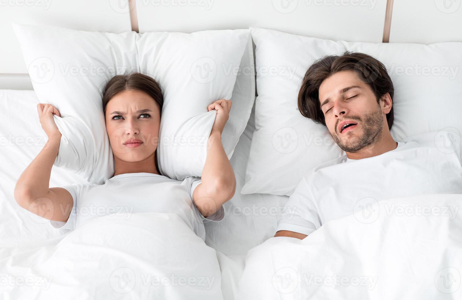 Attractive millennial european guy with stubble sleeps on bed, angry upset wife covers ears with pillow, suffers noise in bedroom interior. Snoring, health and relationship problems, apnea, insomnia photo