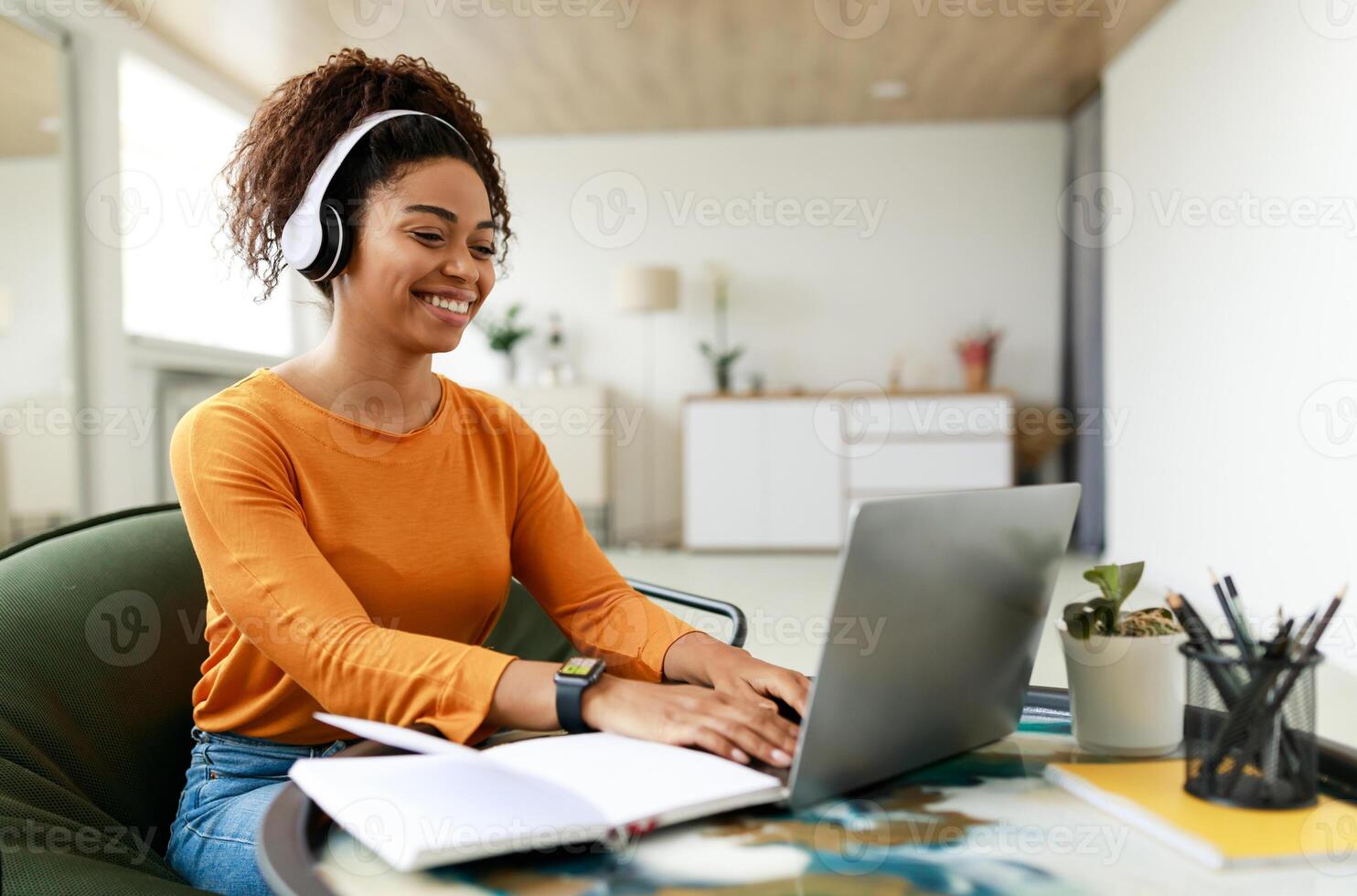 People And Technology. Portrait of young smiling black woman in wireless headset using pc sitting at table on chair in living room, typing on keyboard. Cheerful lady browsing internet, surfing web photo