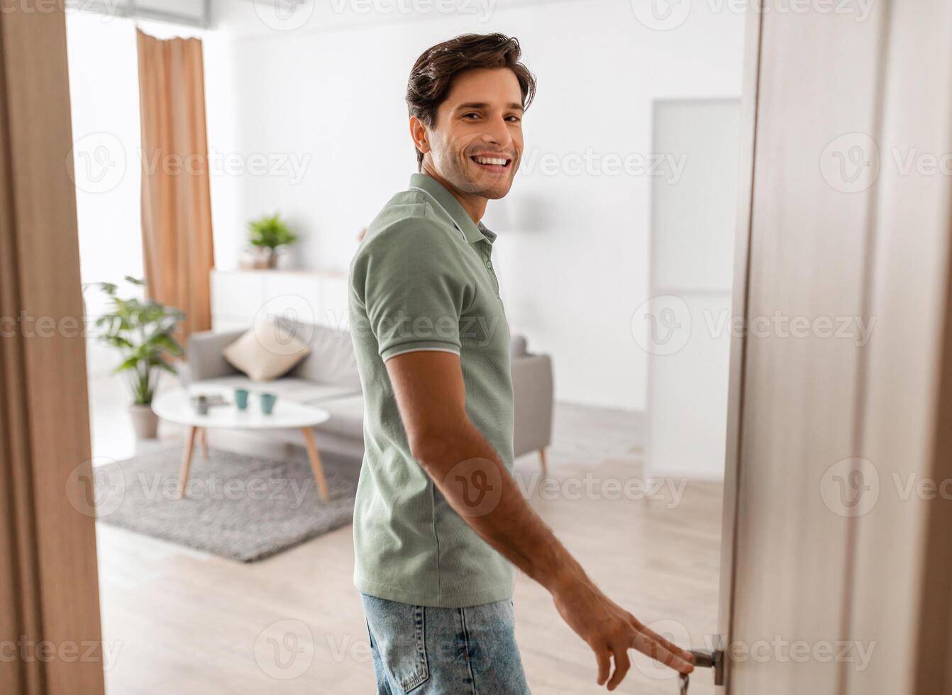 Portrait of excited young man walking in his apartment, entering new home and looking back at camera, happy young guy standing in doorway of modern flat, coming inside, selective focus photo