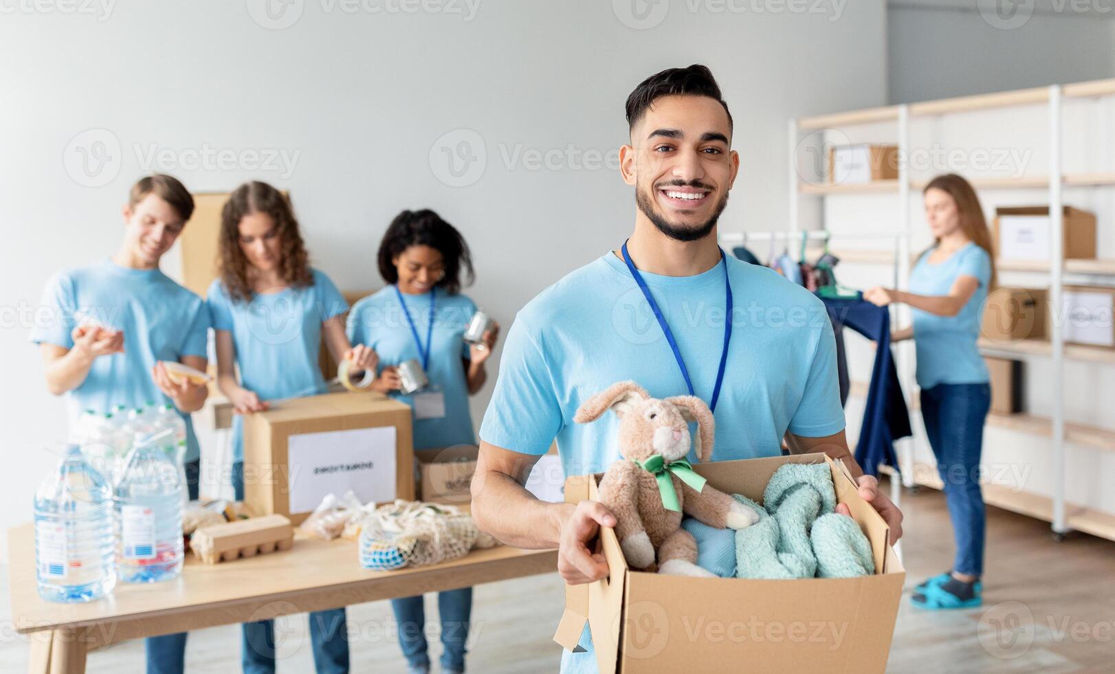 Arab male volunteer holding box with clothes and toys for donation, smiling to camera while working at charity center with group of colleagues, packing products in boxes on background photo