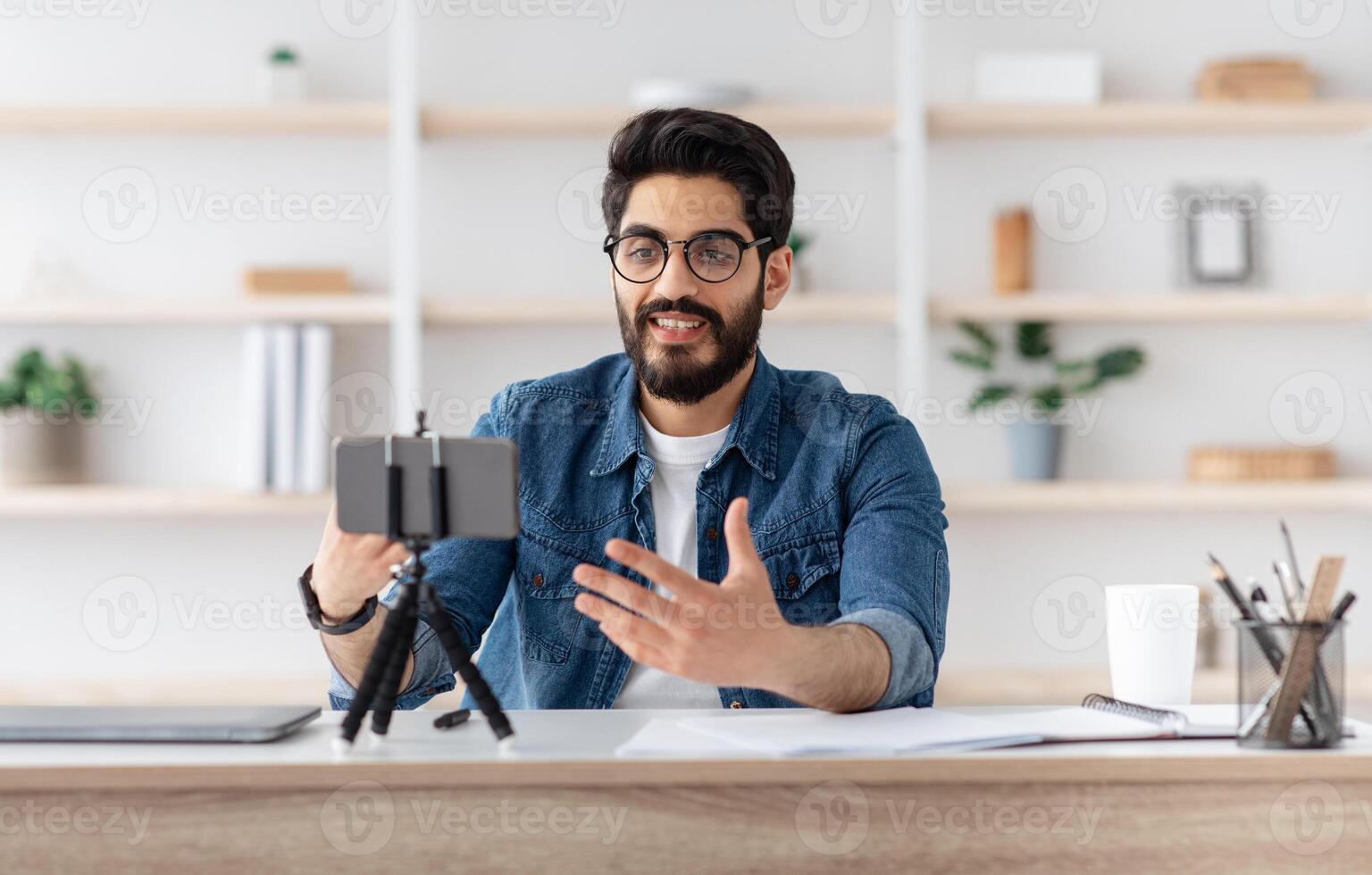 Arab male blogger recording at home with smartphone on tripod, middle-eastern man making content for his blog, sitting at home office and gesturing at phone camera, copy space photo