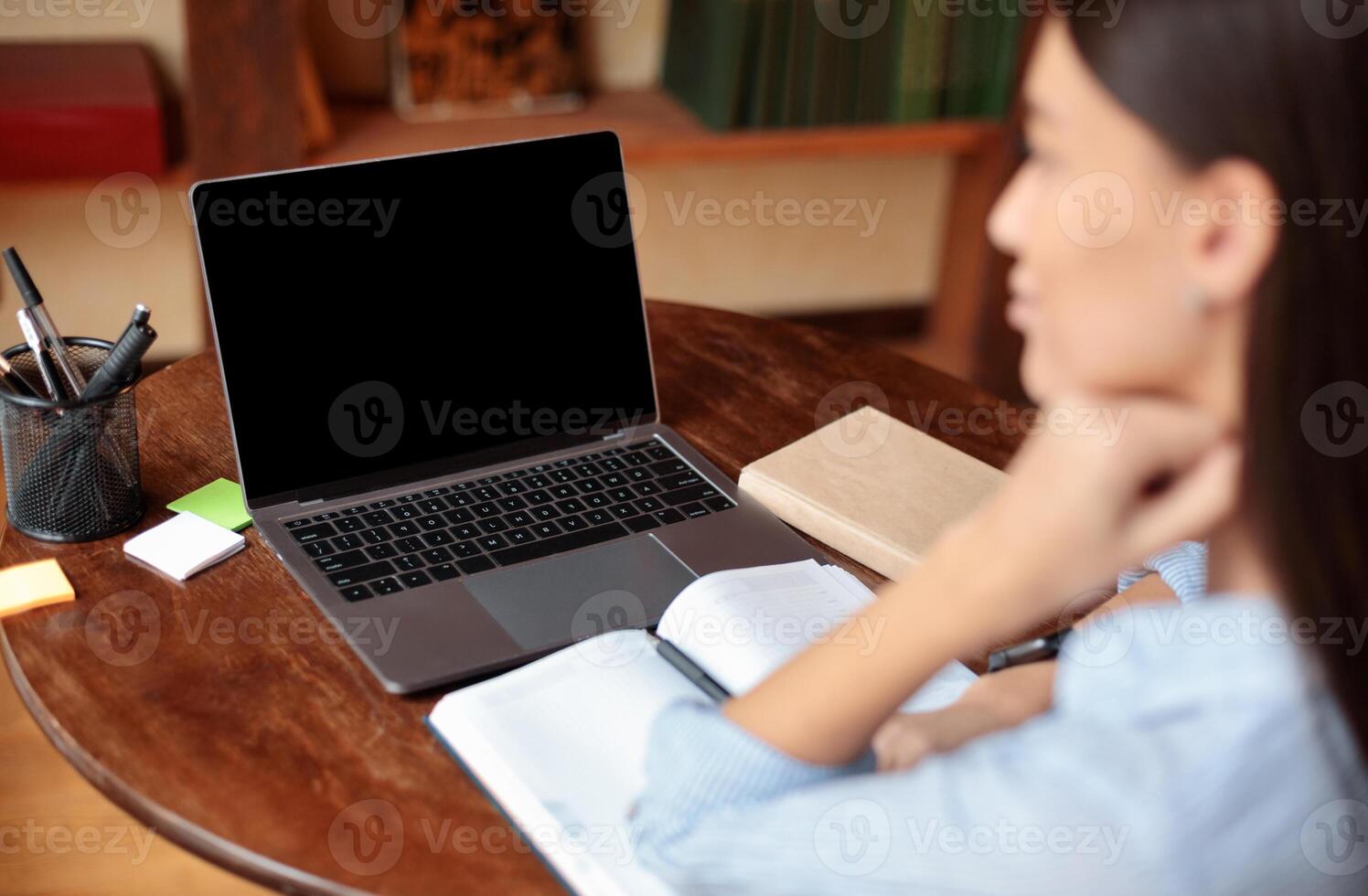 E-learning Concept. Over the shoulder view of woman sitting at desk with open notebook, using laptop with black empty screen for mockup template, watching webinar, studying online at home photo