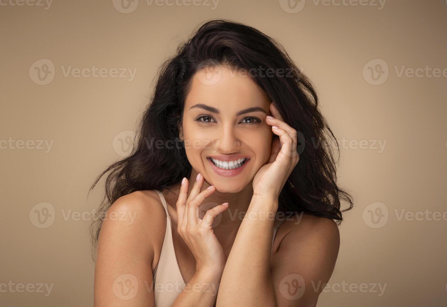 Closeup portrait of sensual beautiful young brunette long-haired woman posing on beige studio background, touching her face and smiling, enjoying her smooth glow face skin after beauty treatment photo