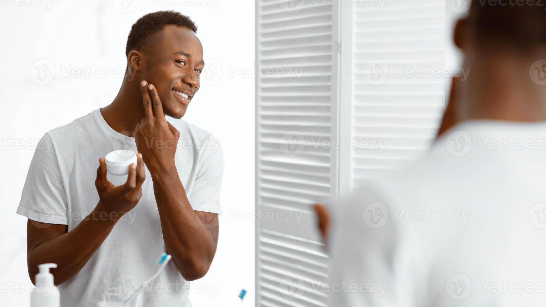African American Man Using Facial Cream Moisturizer Caring For Face Skin Standing Near Mirror In Modern Bathroom Indoors. Male Skincare And Men's Daily Beauty Routine Concept. Panorama photo