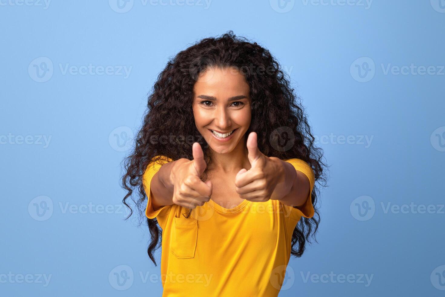 Joyful brunette woman gesturing thumbs up at camera, demonstrating sign of approval, recommending something, positive millennial lady standing over purple background in studio, copy space photo