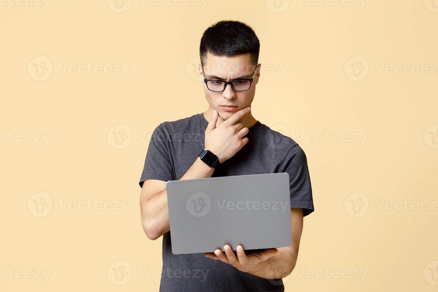 Think about difficult task, online education and work. Pensive busy young man student with glasses presses hand to chin and looks at laptop, isolated on beige background, studio shot, free space photo