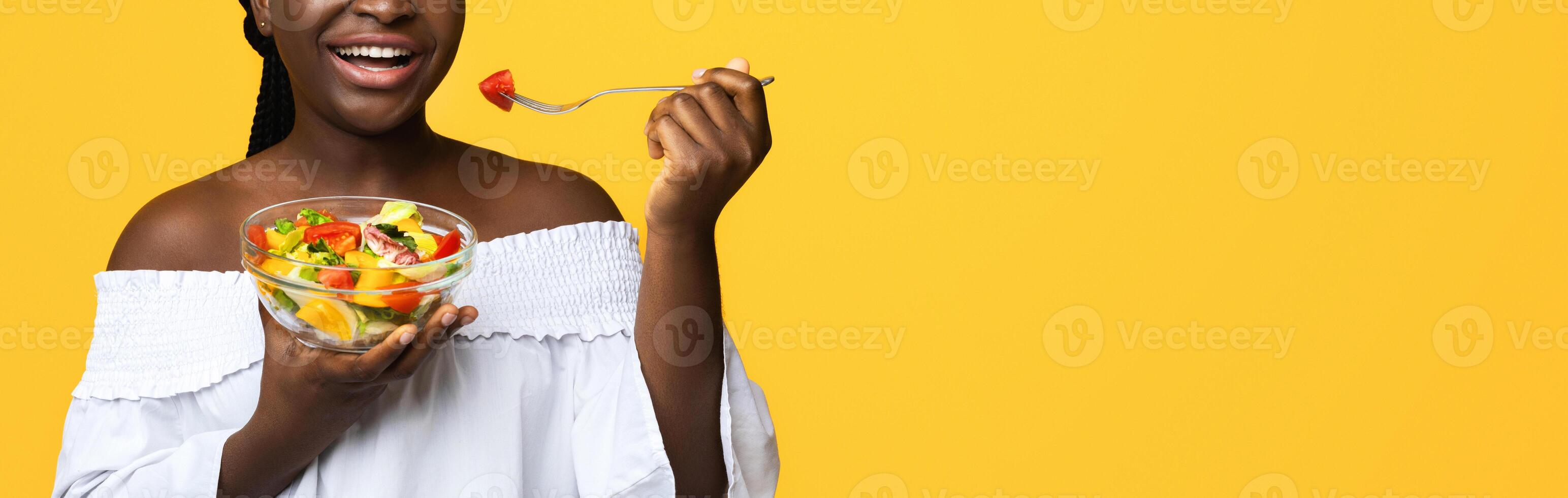 Healthy Nutrition. Happy black young woman eating fresh vegetable salad with fork, holding bowl with vegetarian vitamin food, smiling at camera, isolated over yellow background with copy space photo
