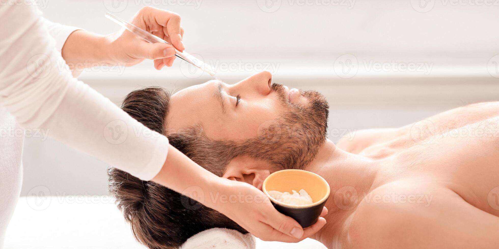 Side view of handsome caucasian middle-aged man getting face treatment at spa, closeup. Unrecognizable beautician applying detox white mask on relaxed man face, using brush and clay photo