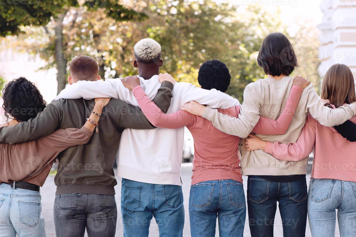 Back view of international group of people in casual outfits hugging on the street, unrecognizable multiracial students standing in a row and embracing outdoors, panorama photo