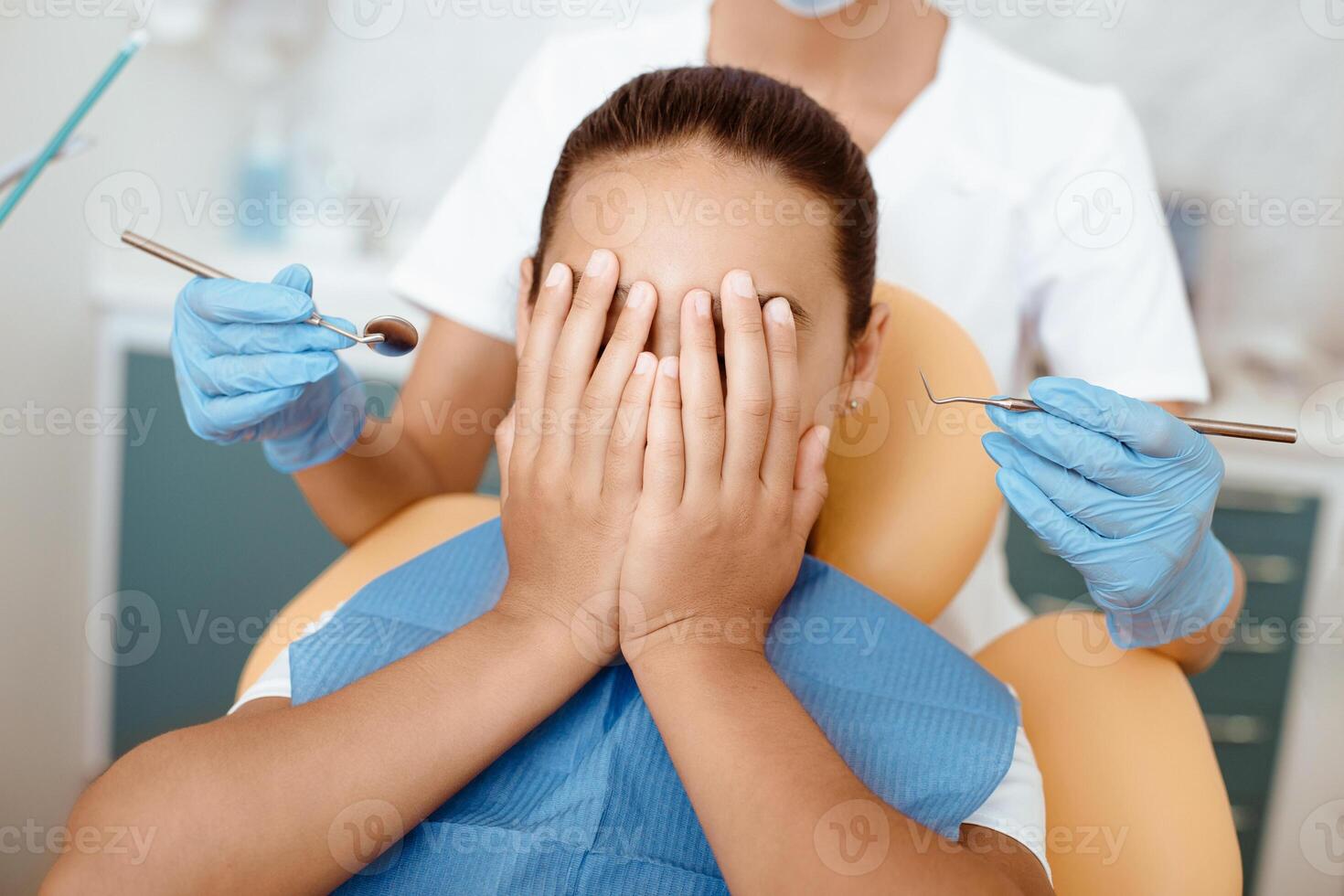 Modern dental treatment and fear. Scared girl covers face with hands and does not allow doctor in white uniform and rubber gloves to treat mouth with medical instruments in hands, cropped, close up photo
