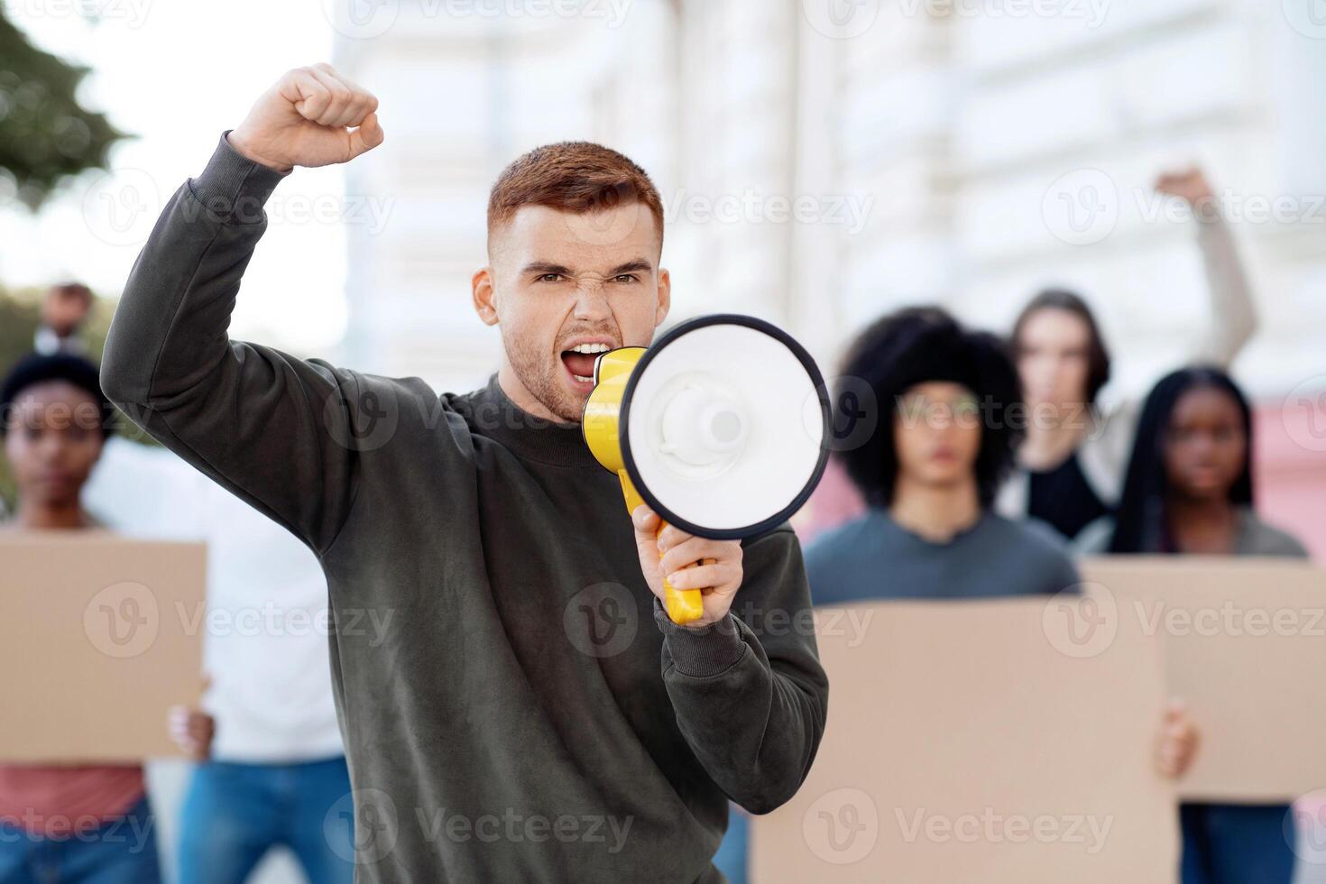 Young man with megaphone leading international group of protestors, walking by street together, holding empty placards and chanting slogans. Multiracial group of students defending human rights photo