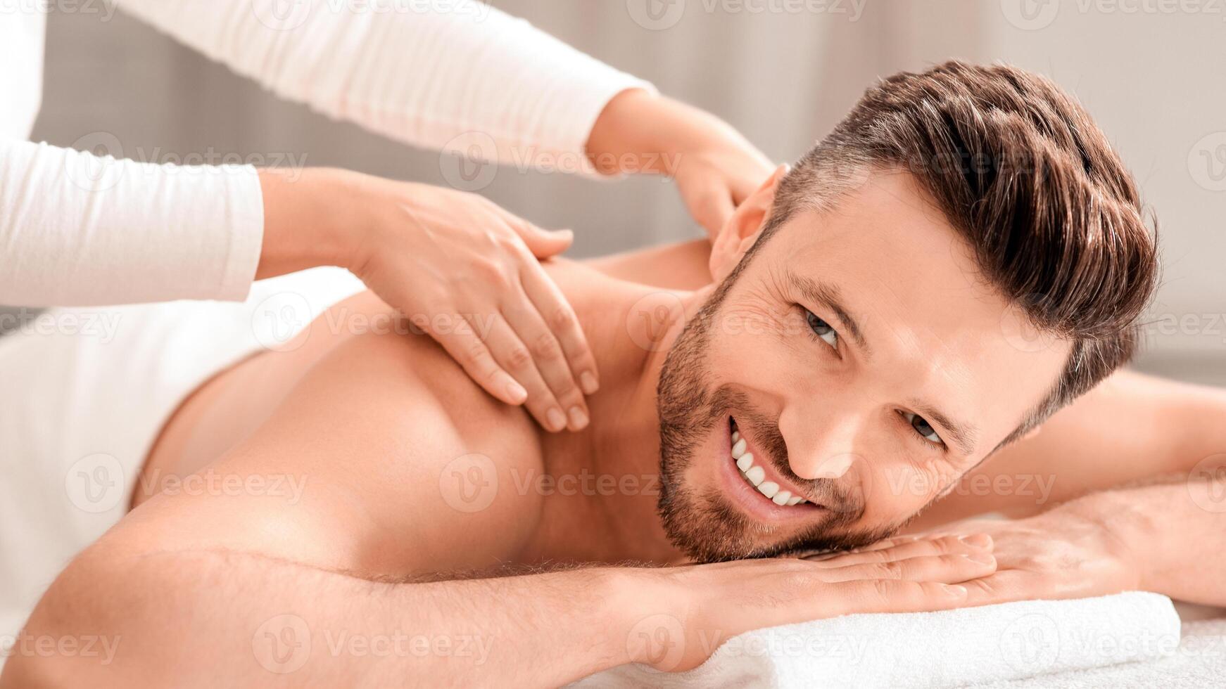 Cheerful middle aged man attending spa salon on weekend, closeup. Handsome bearded man smiling at camera while getting relaxing back massage. Female therapist rubbing joyful man shoulders at spa salon photo