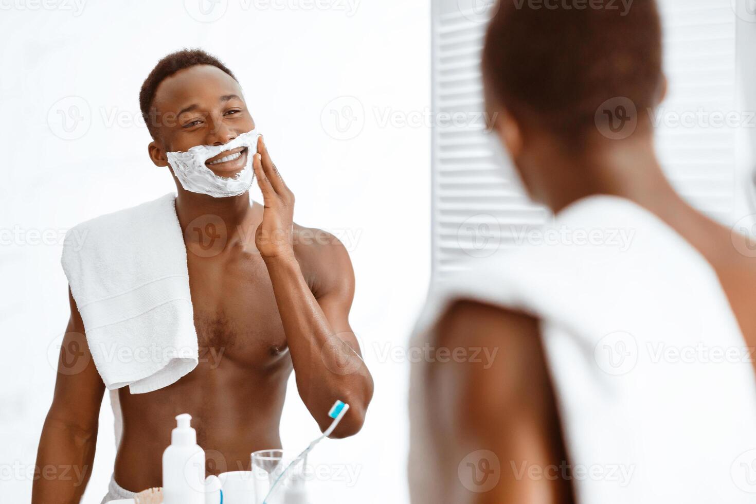 Shirtless Black Man Preparing For Shaving Foaming Face Applying Shave Foam Standing Near Mirror In Modern Bathroom At Home. Male Selfcare And Facial Skincare Routine. Selective Focus photo