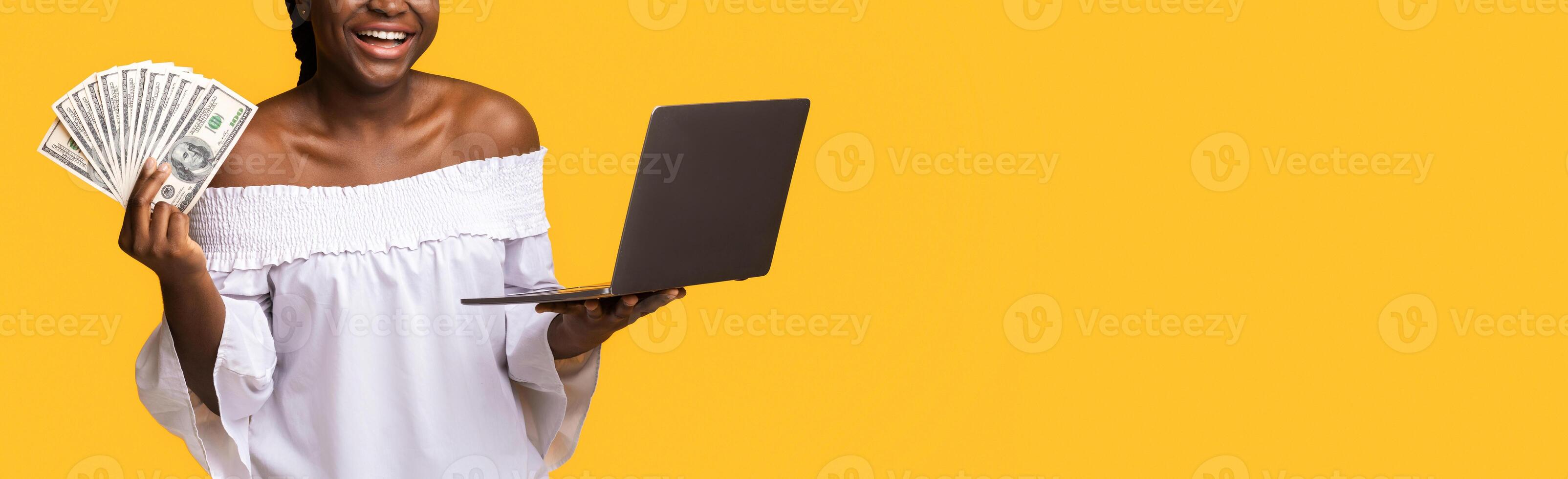Making money online. Excited african american woman holding dollar cash and laptop, enjoying e-commerce profit, won lottery, working in internet, posing over yellow studio background with copy space photo