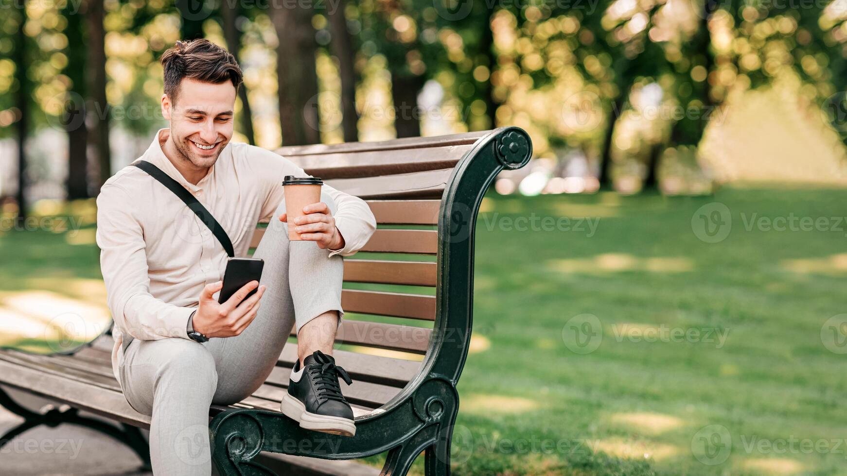 A young man relaxes on a bench in a vibrant urban park, sipping coffee from a cup and looking at his smartphone. The sun shines through the trees, creating a lively atmosphere, copy space photo