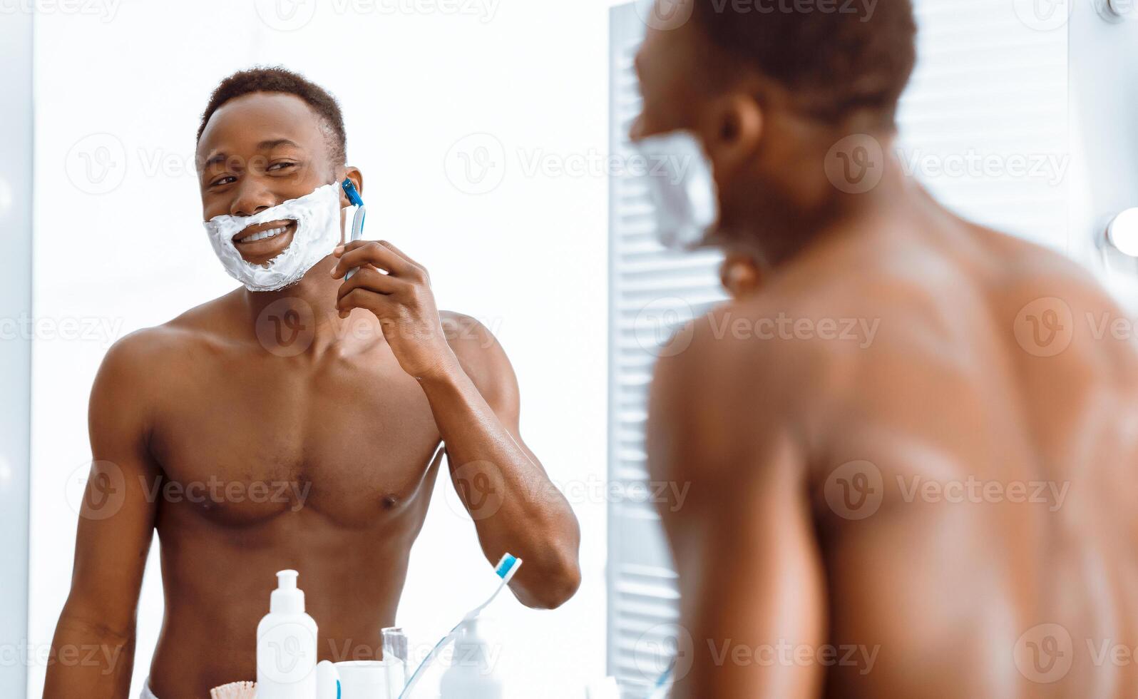 Handsome Shirtless Black Man Shaving His Face With Razor And Shave Foam Standing In Modern Bathroom In The Morning. Male Beauty Routine And Smooth Skin Concept. Selective Focus photo