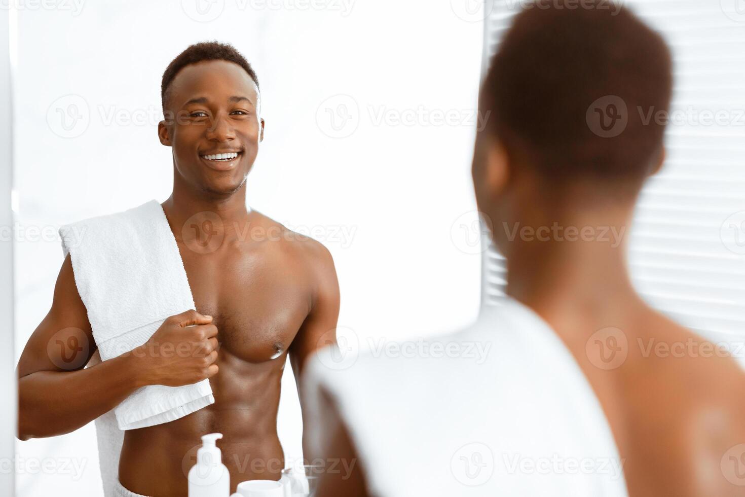 Attractive Shirtless Muscular African Man Holding Towel Standing In Modern Bathroom At Home, Looking In Mirror At His Reflection. Male Self Care, Morning Beauty Routine Concept. Selective Focus photo