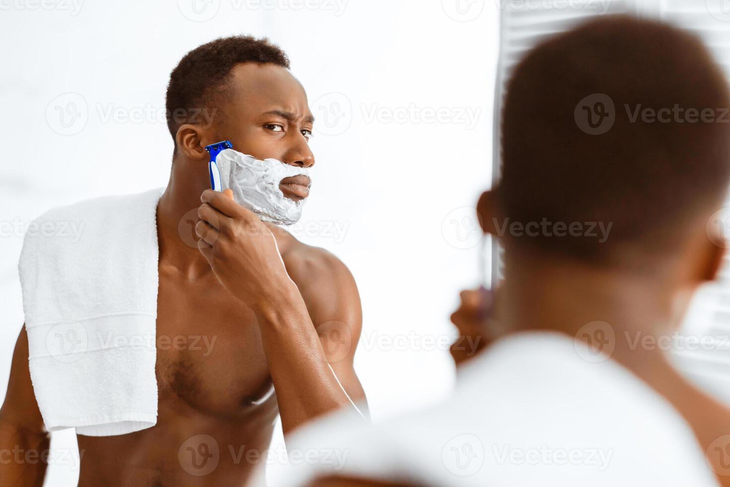 African American Man Shaving Face Covered With Shave Foam Standing Shirtless Near Mirror In Modern Bathroom At Home In The Morning. Male Selfcare Beauty Routine And Facial Skincare. Selective Focus photo
