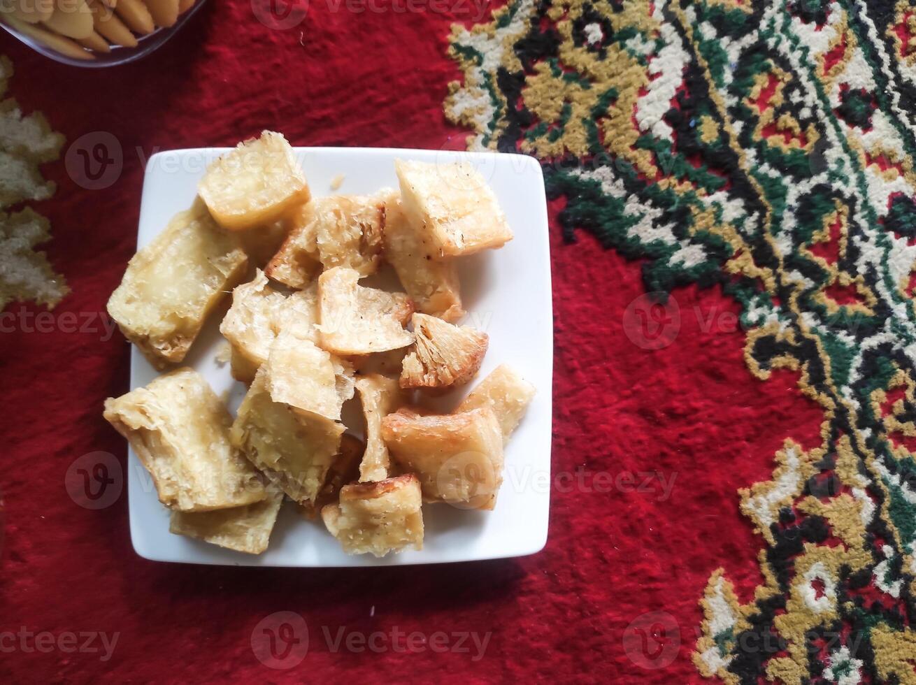 Photo of a plate of fried cassava. Fried food served on a white plate on a red Persian carpet. Traditional Indonesian cuisine. Soft and crunchy texture. Tasty Gourmet. Text space and closeup