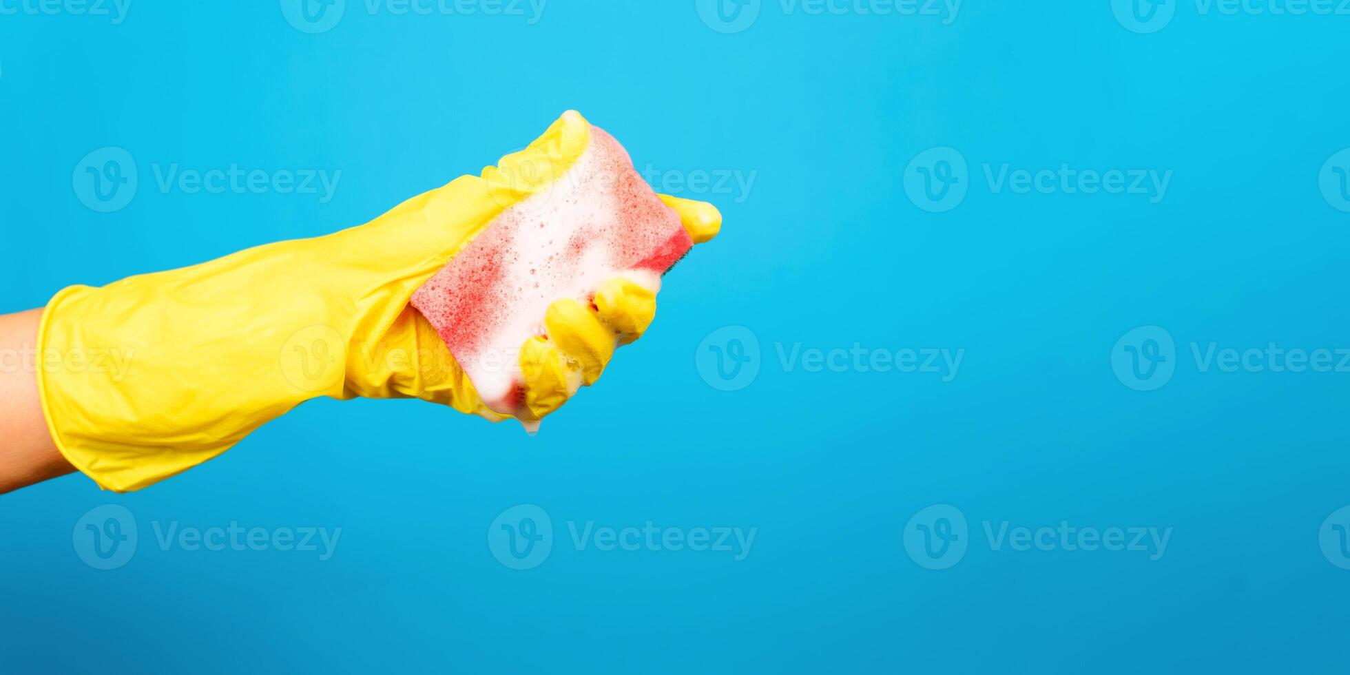 Female hand in yellow glove squeezing foamy sponge for washing. Blue background, copy space. House cleaning, washing concept photo