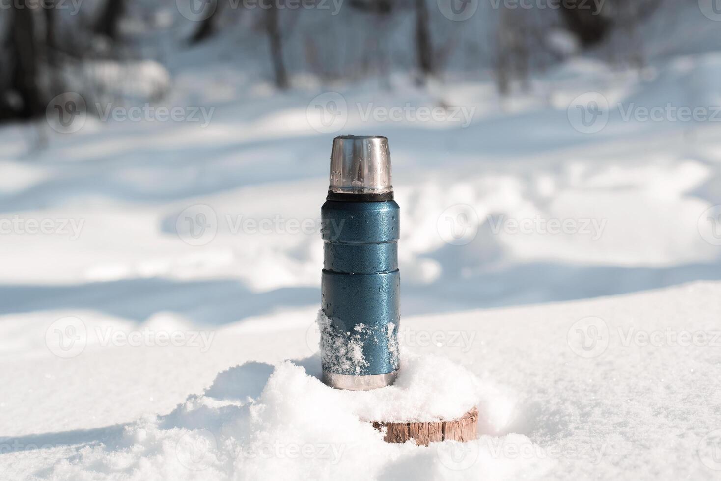 Metal blue thermos standing on a snowy tree stump in a winter forest on a sunny day photo