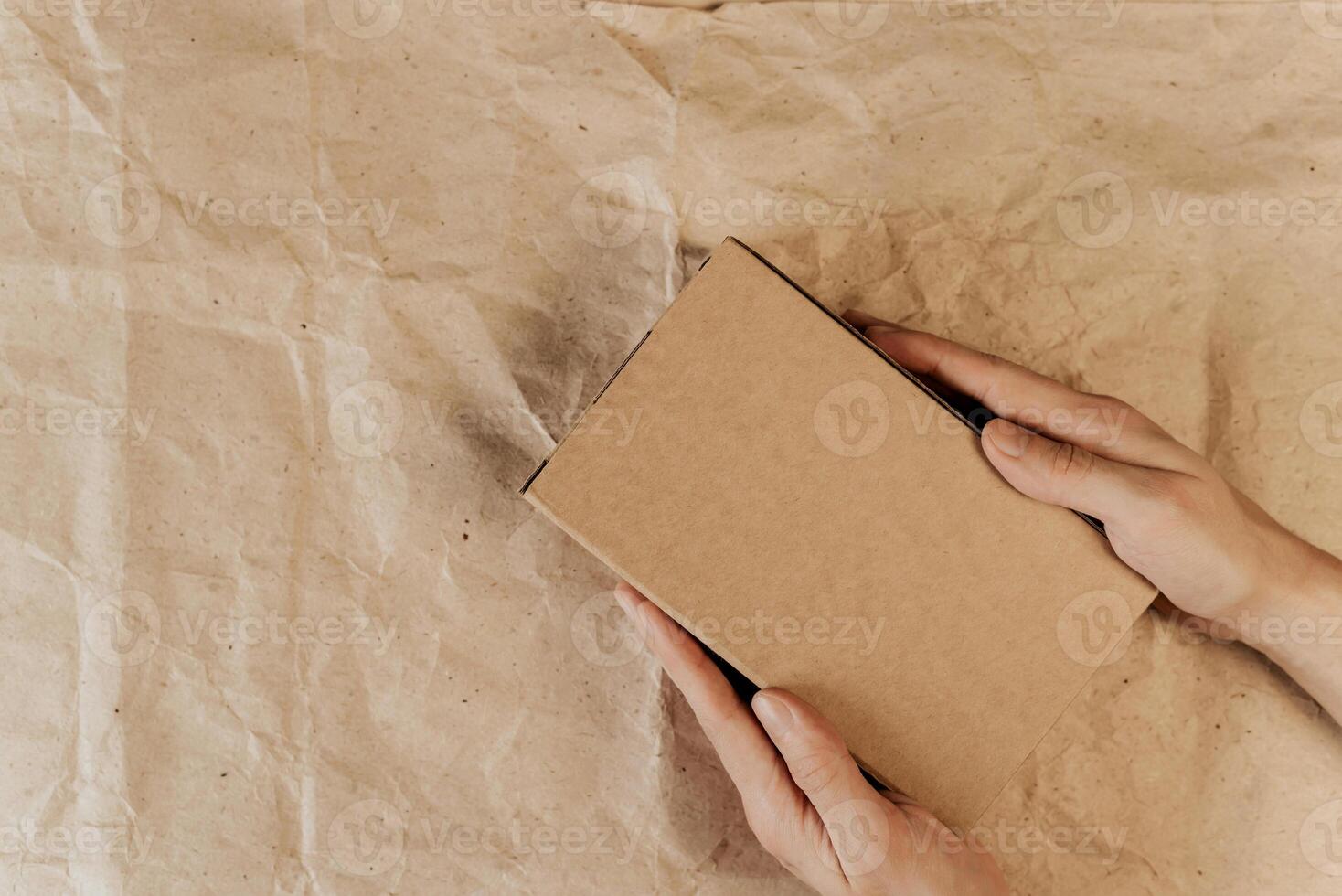 Hands holding brown cardboard box, wrapping paper background. Top view, layout. Place for logo, announcement, text. Gift, delivery concept photo
