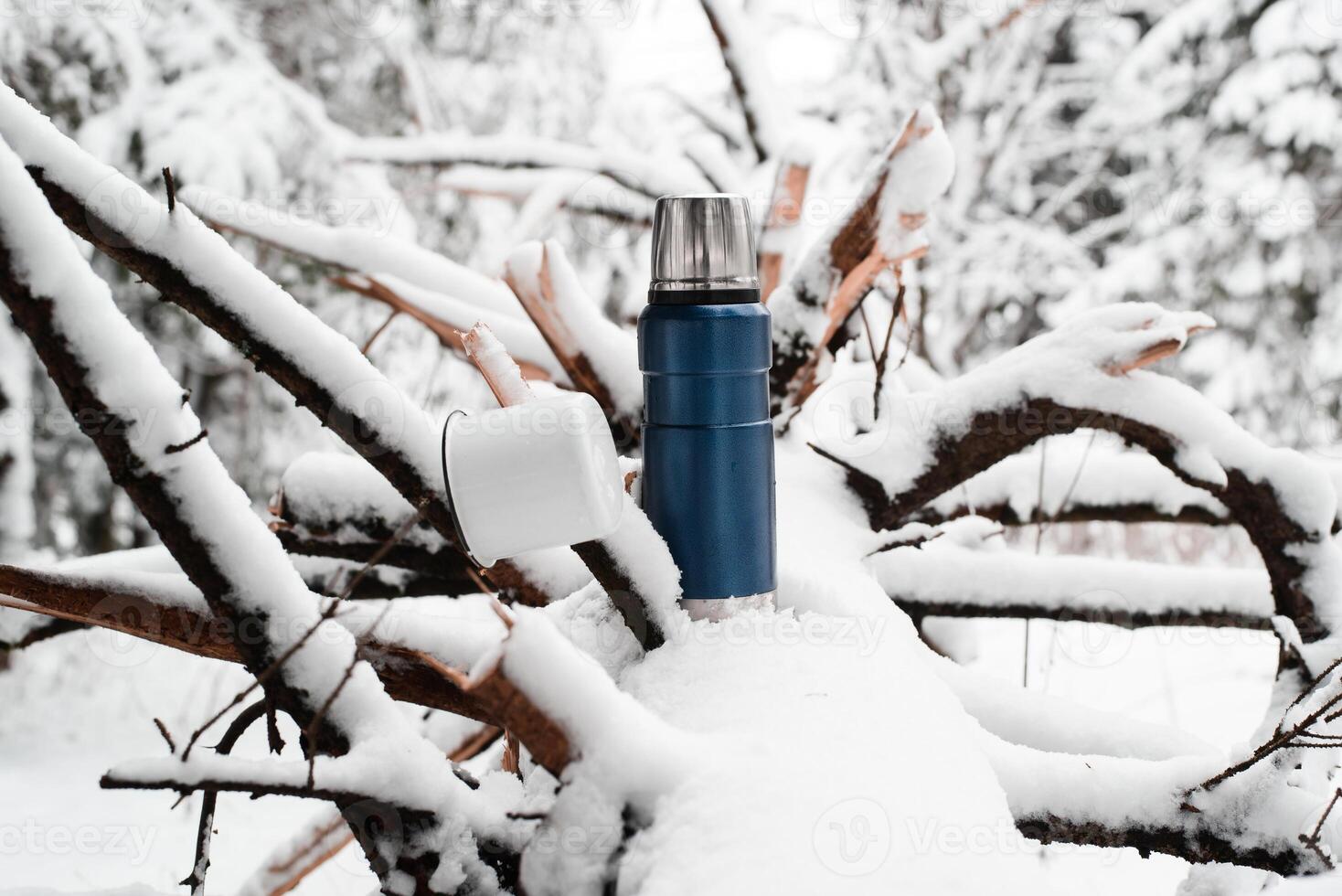 Thermos and an iron mug on a fallen pine tree. Camping in the winter forest. Snow covered trees. Vacuum Travel Flask photo